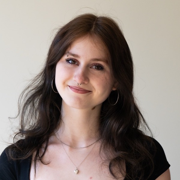 A portrait of Penelope Suchar, a person with dark brown wavy hair, wearing a black scoop-neck top, hoop earrings, and a pendant necklace.