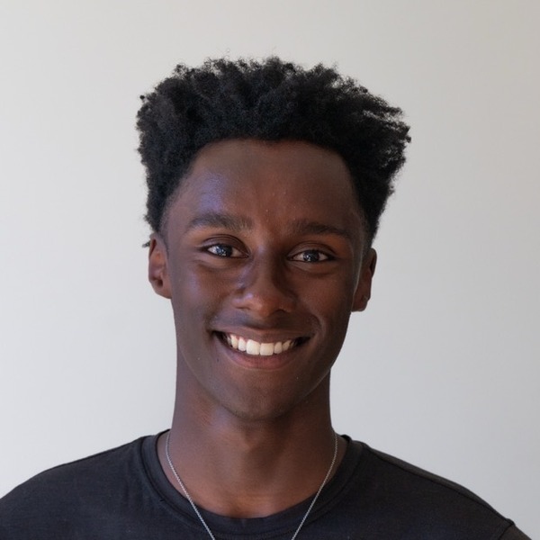 A portrait of Sam Stableford, a person with black hair, wearing a charcoal crew-neck shirt and layered necklaces.