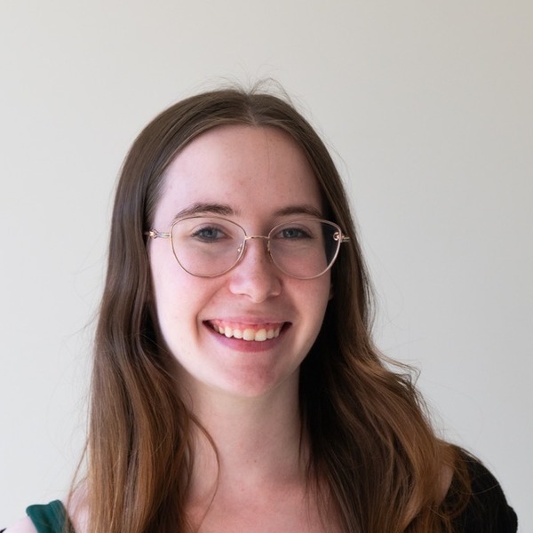 A portrait of Stella Johnson, a person with long light brown hair, wearing glasses and a black off-the-shoulder top.