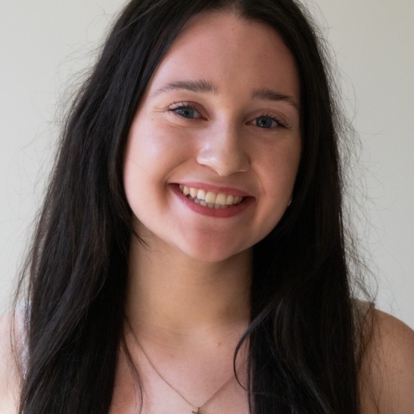 A portrait of Tara Sweeney, a person with long dark brown hair, wearing a white sleeveless top and a pendant necklace.