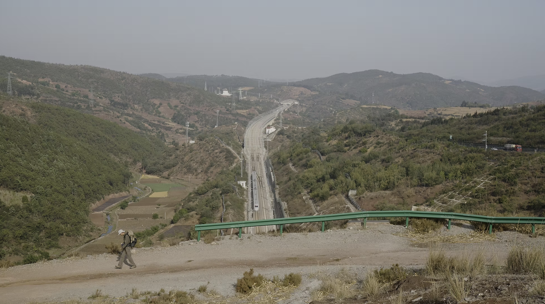 A person with a backpack walks along a hillside road overlooking railway tracks stretching through a valley of wooded hills and farmland.