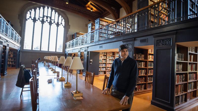 Ronald Patkus standing at a table in the Vassar Library with book shelves behind him.