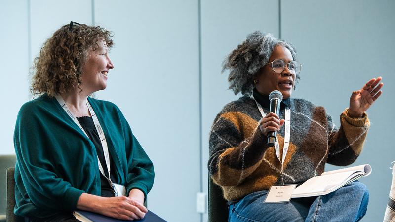 Two people sitting on a stage with one speaking into a microphone to an audience.