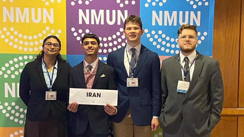 A group of smiling people standing in front of a backdrop with the acronym NMUN repeated: . A person in the middle is holding a sign that reads: Iran.