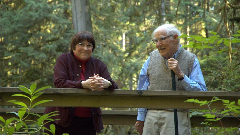 An elderly man and woman standing together on a wooden walkway in a lush, forested area, smiling and talking.
