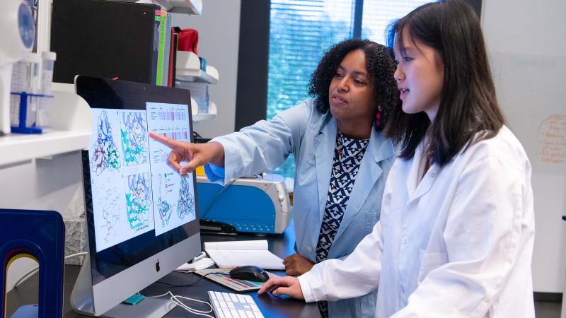 Two people wearing lab coats examine 3D protein structure models on a large computer monitor in a laboratory. One person points at the screen while the other operates the computer.