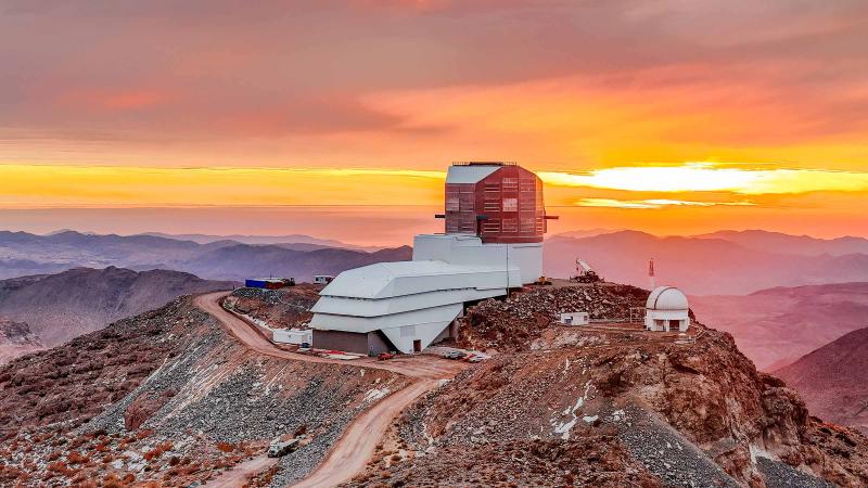 Vera C. Rubin Observatory sits atop a rocky mountain ridge at sunset, with vibrant orange and yellow hues lighting the sky.