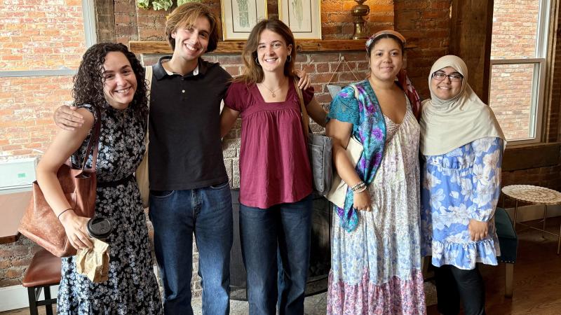 A professor and four students, all smiling, pose in front of a brick fireplace.