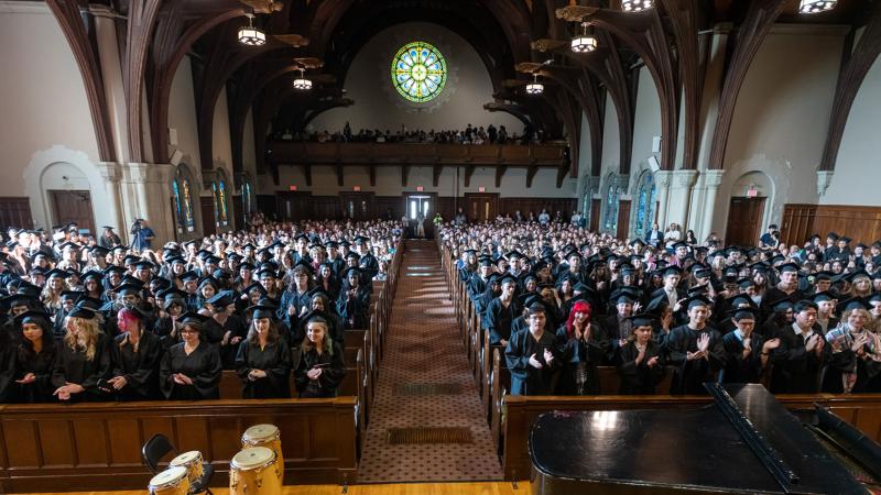 Large group of graduates in black caps and gowns filling a chapel, standing and applauding during a commencement ceremony, with stained glass windows and wooden beams visible overhead.
