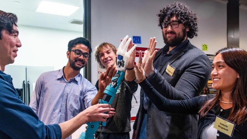 A group of onlookers checking out a robotic hand, held by a person, offerring a high five.