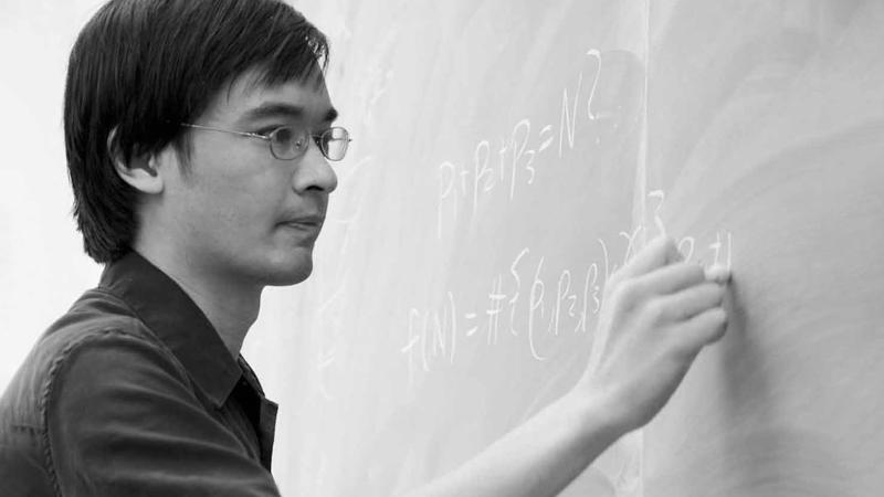  Black-and-white photo of a person writing mathematical formulas on a chalkboard.