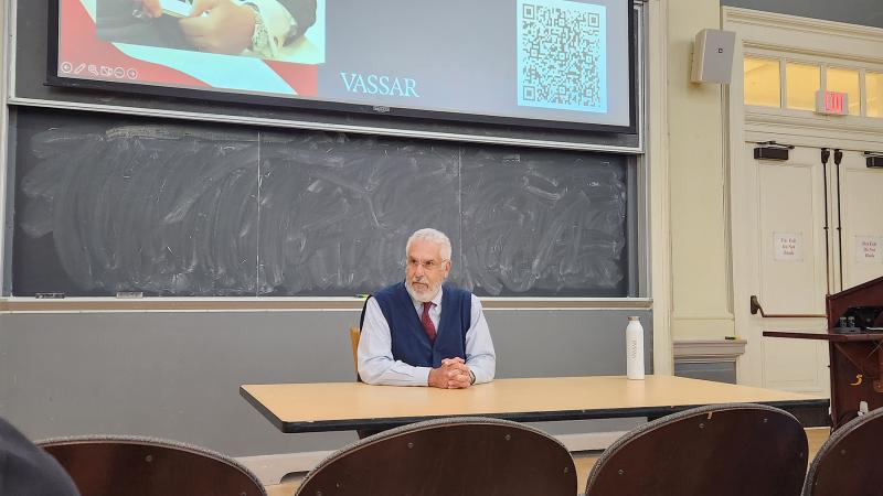 Person seated at a classroom table in front of a chalkboard, with a Vassar presentation slide and QR code projected above.