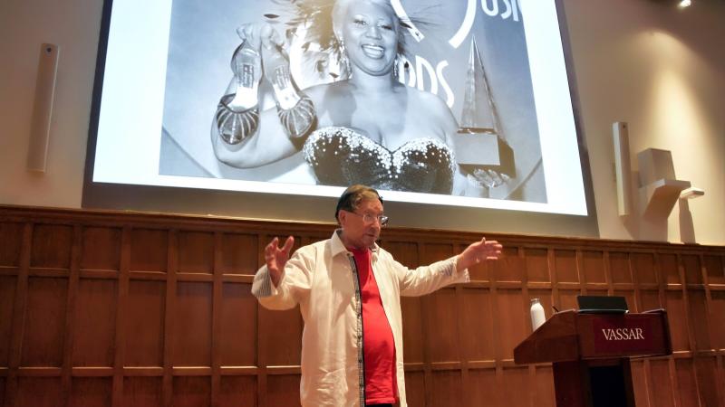 A person standing and speaking before and audience and in front of a large projection screen with their arms spread wide.