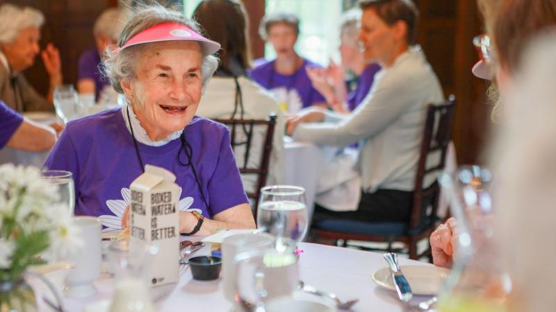 June Ross Marks ’49 with short white hair wearing a bright purple shirt and a pink visor, seated at a dining table.
