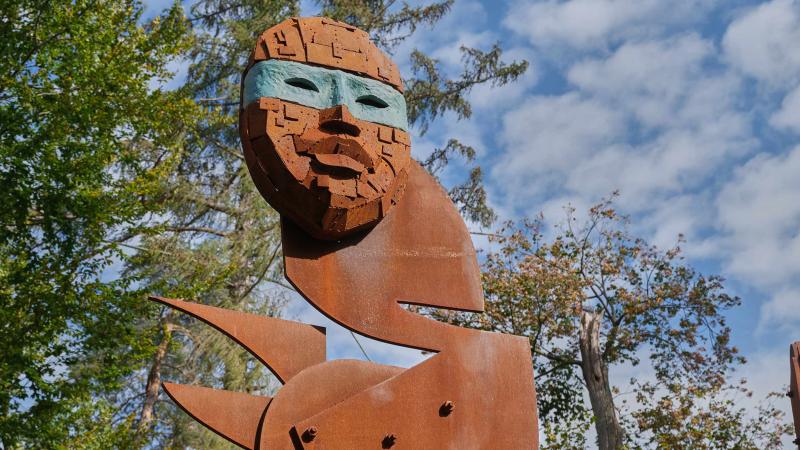  A tall outdoor sculpture with a weathered, rusted metal face and body. The mask-like face has an abstract, blocky design and is accented by a smooth, teal-colored band over the eyes. Trees and a blue sky with white clouds form the background.