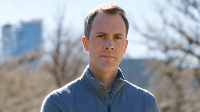 Outdoor head-and-shoulders portrait of a person in a gray quarter-zip, neutral expression, with blurred trees and a blue building behind the person.