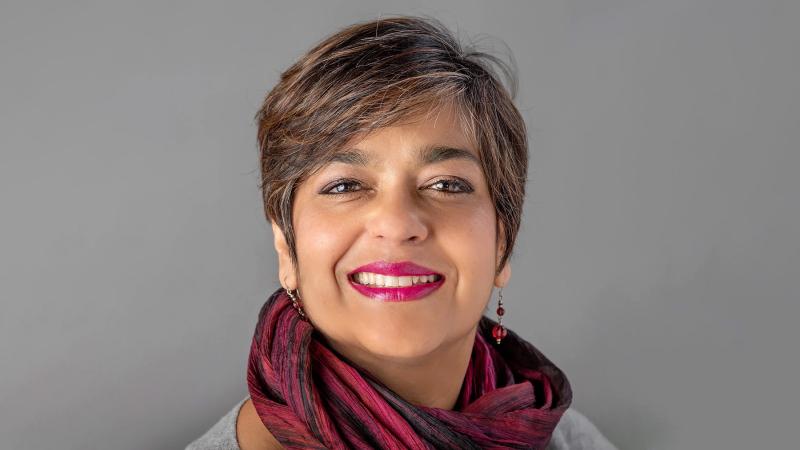 Smiling woman with short dark hair, fuchsia lipstick, and a burgundy scarf, posed against a plain gray background.