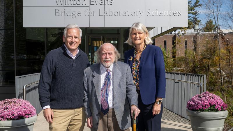 Three adults stand smiling in front of a campus building sign that reads “Winton Evans Bridge for Laboratory Sciences”; the man in the center holds a cane, pink mums in planters flank the walkway, and fall trees are visible in the background.