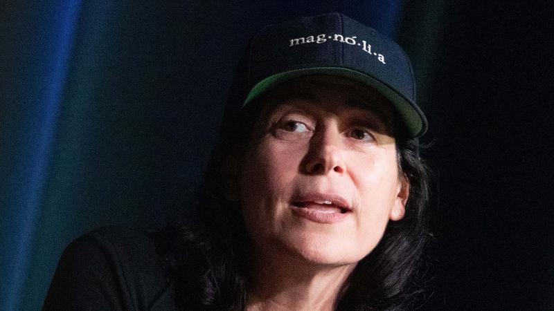 A person wearing a dark shirt and a "mag·no·li·a" baseball cap speaks into a microphone while gesturing with their right hand.