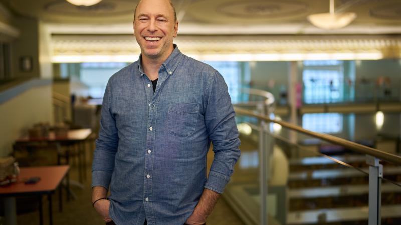 A smiling person wearing a denim shirt stands with their hands in their pants pockets in an indoor atrium with big windows.