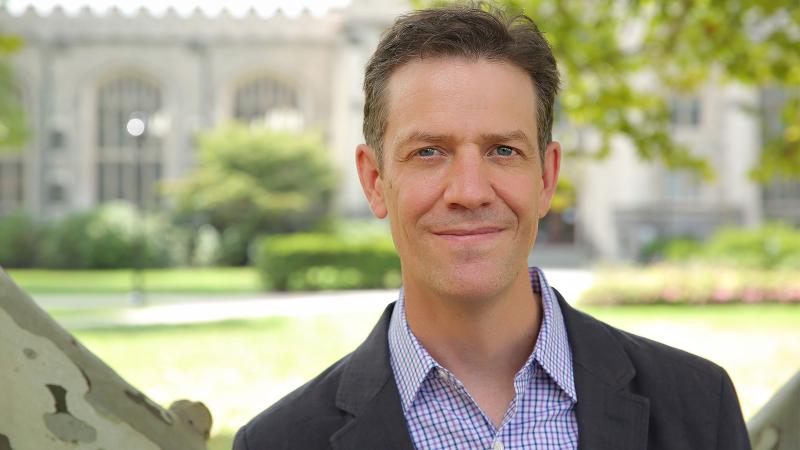 A smiling person wearing a collared shirt and blazer stands on the Vassar campus with the college library in the background.