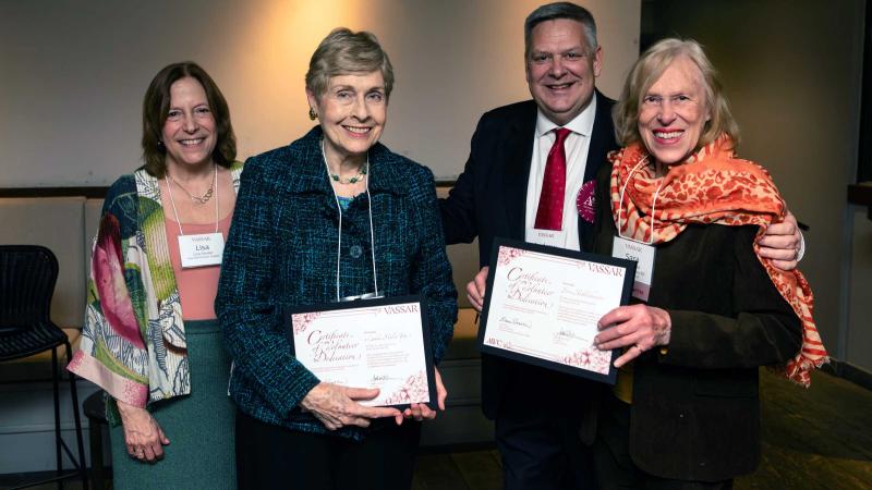 Four smiling people standing side by side while two in the center hold framed documents.