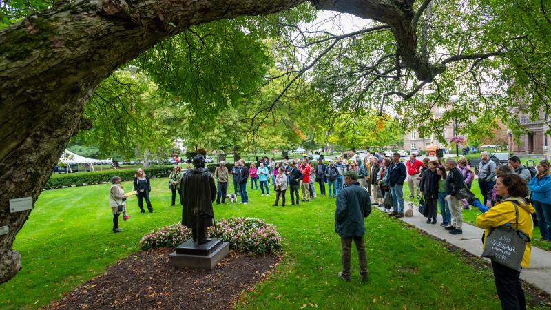 Group of visitors gathered on a campus lawn around a statue during an outdoor tour, with large tree branches overhead.