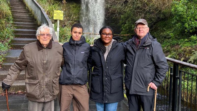 Four people stand outdoors, with foliage and a stone staircase behind them.