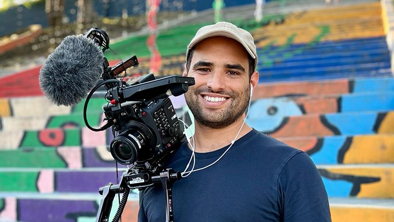 A smiling man stands beside a video camera and microphone, with a colorful mural behind him.