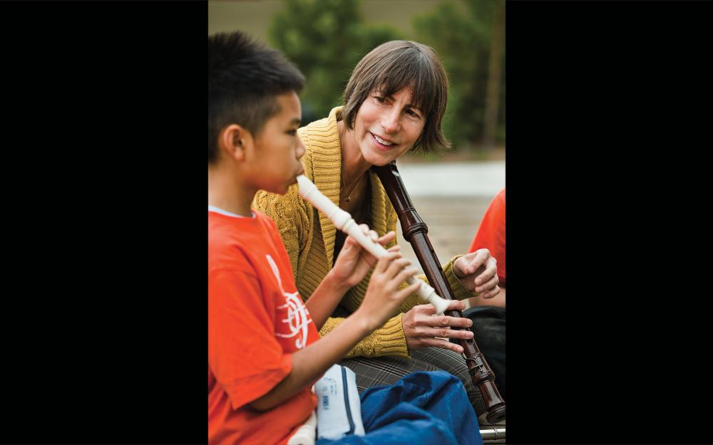 A person, with short dark hair wearing a yellow sweater holding an instrument, faces a child with short dark hair wearing a bright orange shirt playing the flute.