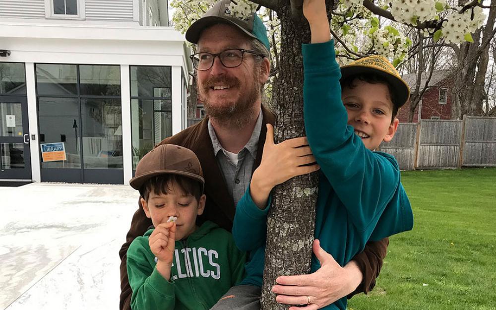 A person with light hair wearing a baseball cap and a brown jacket holding onto a tree with his two young children in his arms.