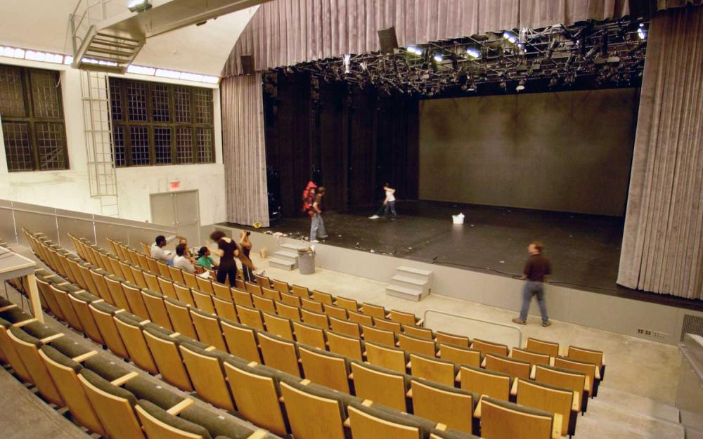 A mostly empty theater, with rows of seats. On stage, a rehearsal is taking place.