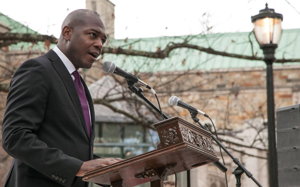 A person with a bald head wearing a formal black jacket and purple tie speaks into a microphone behind a wooden podium.