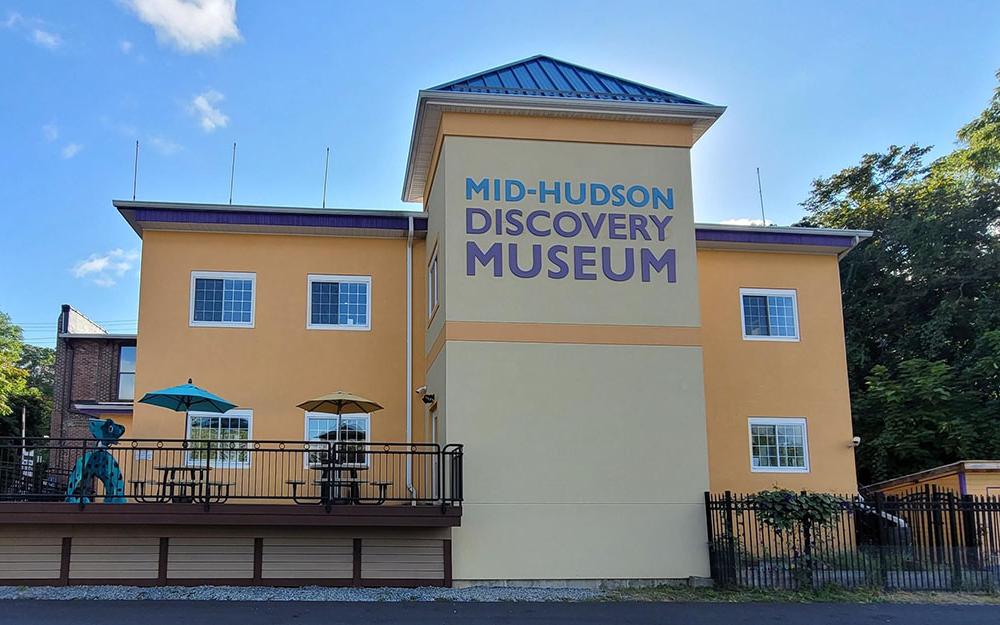 A yellow 2 story building with a sign for the Mid-Hudson Discovery Museum and a few picnic tables with umbrellas outside
