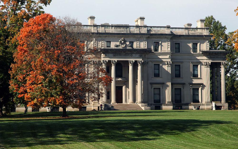 The mansion is Beaux-Arts architecture style painted white with large columns around the entrance on a large green lawn surrounded by trees