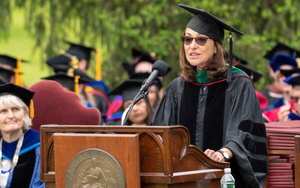 Commencement speaker speaking behind a podium and microphone with faculty and administrators watching