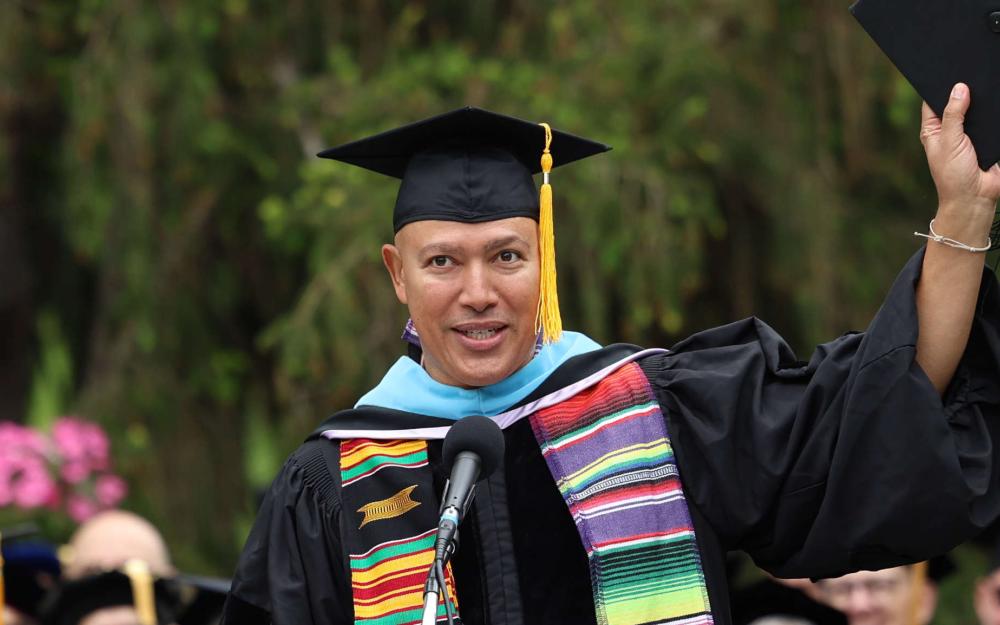 A speaker wearing a black graduation gown, colorful stole, and light blue hood raises a mortarboard in one hand while standing at a microphone. The podium bears the Vassar College seal, and seated individuals in caps and gowns are visible behind. The background includes green trees and clusters of pink flowers. A stack of red diploma covers sits on the stage nearby.