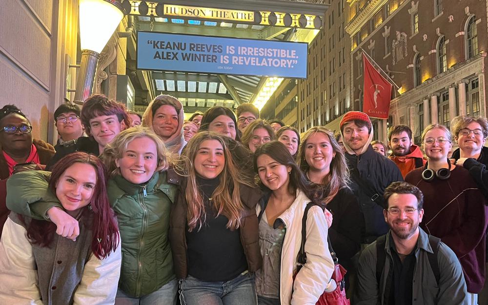 A large group of smiling students poses for a nighttime photo on a city sidewalk outside the Hudson Theatre, with the glowing marquee above them reading, “Keanu Reeves is irresistible, Alex Winter is revelatory."