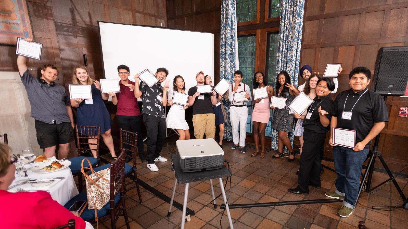A large group of people holding up certificates in a large wood-adorned room with a projector screen in the background.