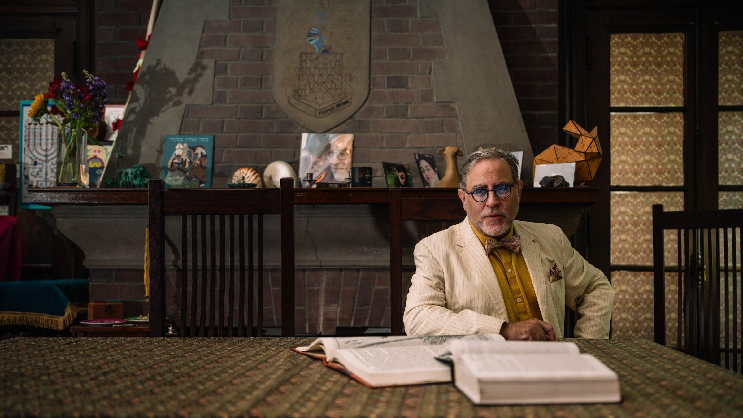 Person in a white suit seated behind a table behind two open books.