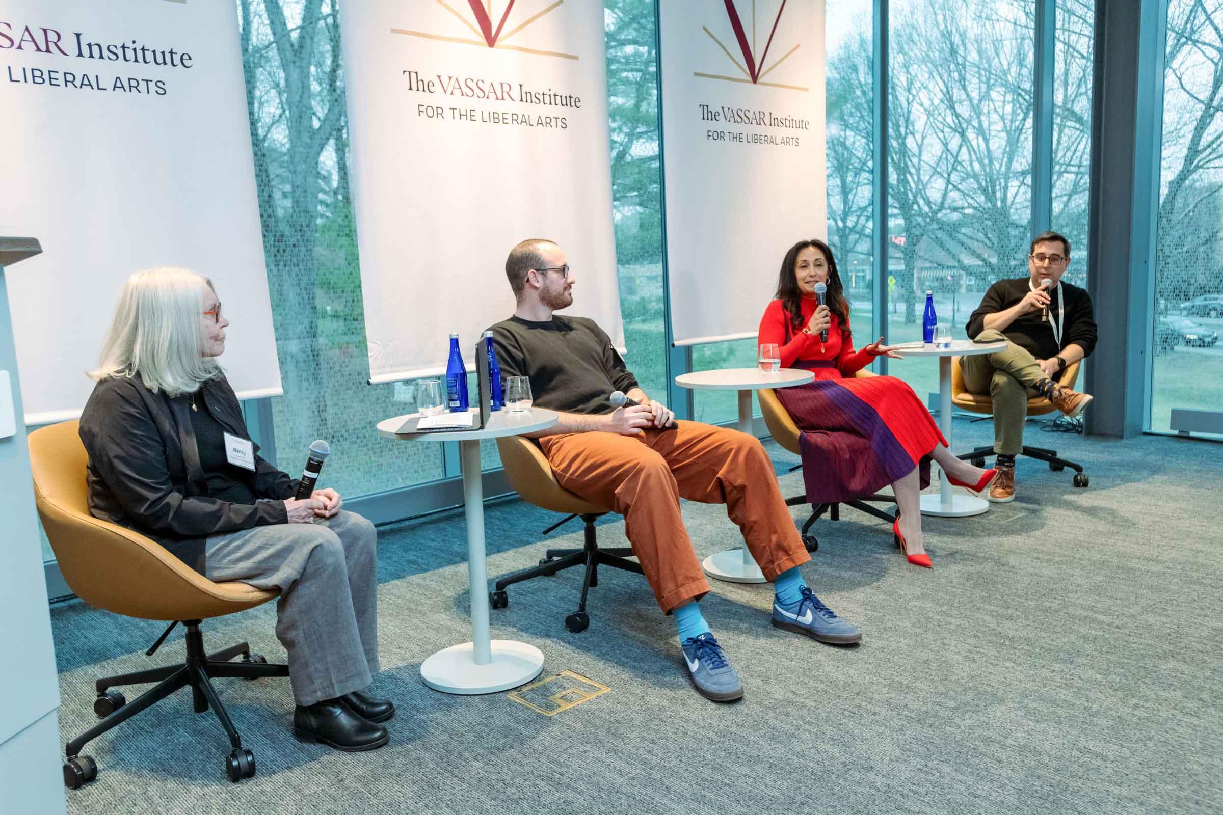 A seated panel of four people listen while one of the panelists holds and speaks into a microphone.