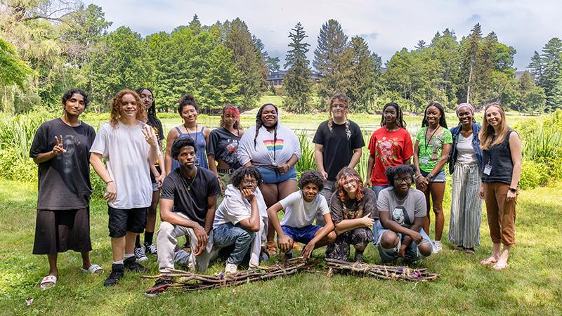 A diverse group of fifteen people is posing for a photo on a grassy lawn with a pond and a college building visible in the background.