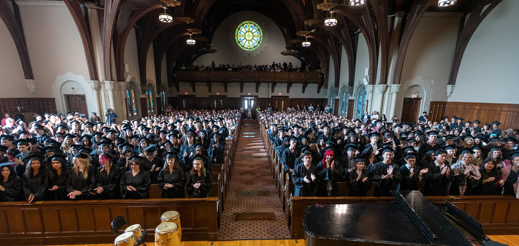 Large group of graduates in black caps and gowns filling a chapel, standing and applauding during a commencement ceremony, with stained glass windows and wooden beams visible overhead.