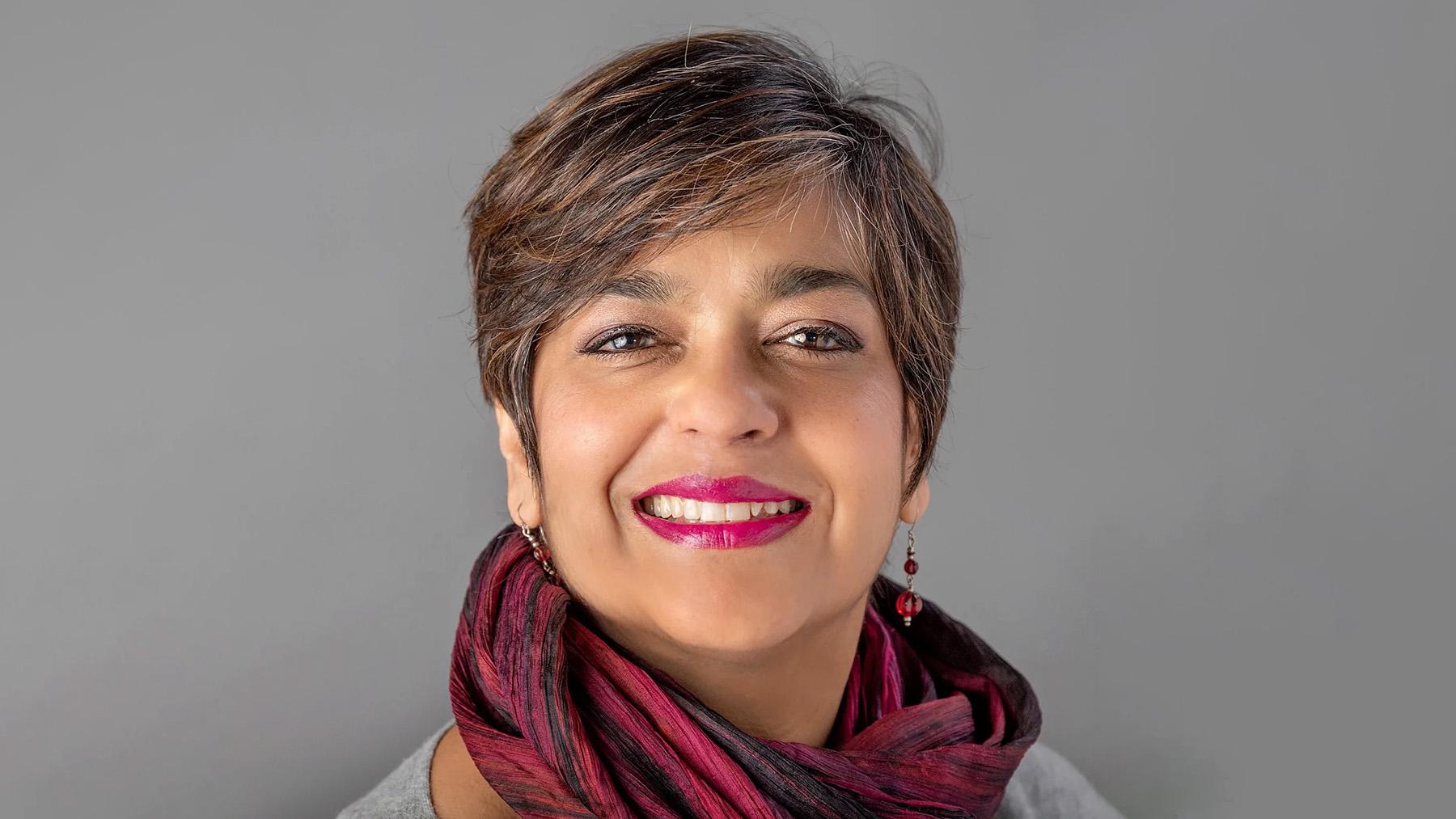 Smiling woman with short dark hair, fuchsia lipstick, and a burgundy scarf, posed against a plain gray background.