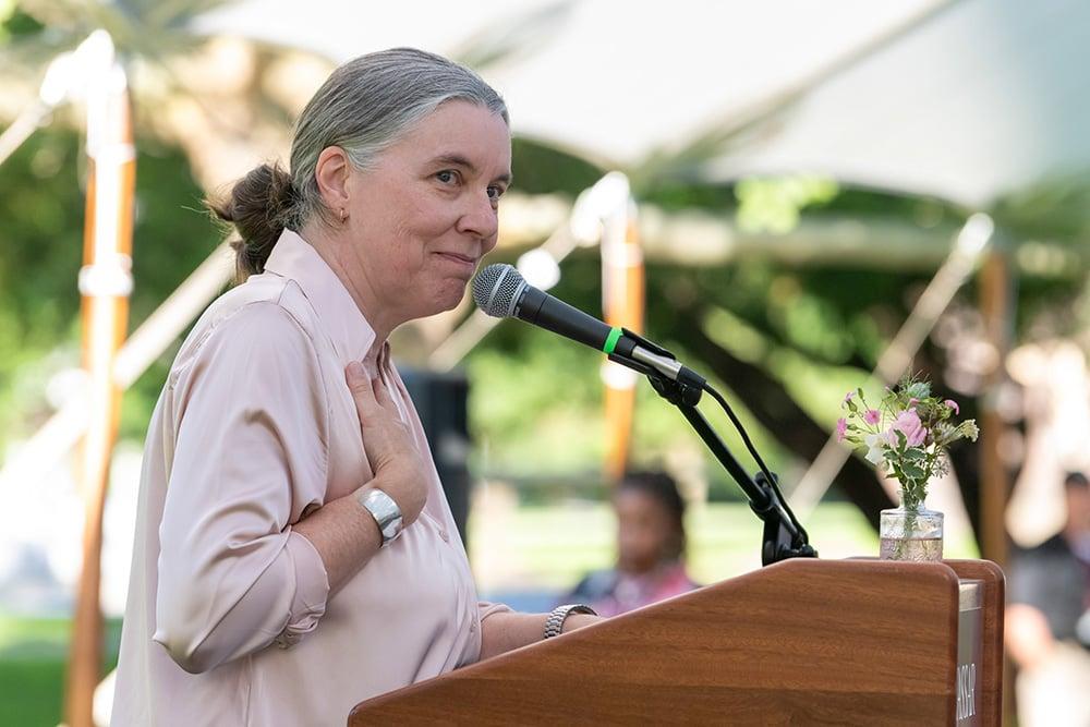 A person with grey hair pulled back in a bun wearing a pink shirt speaks at an outdoor podium.