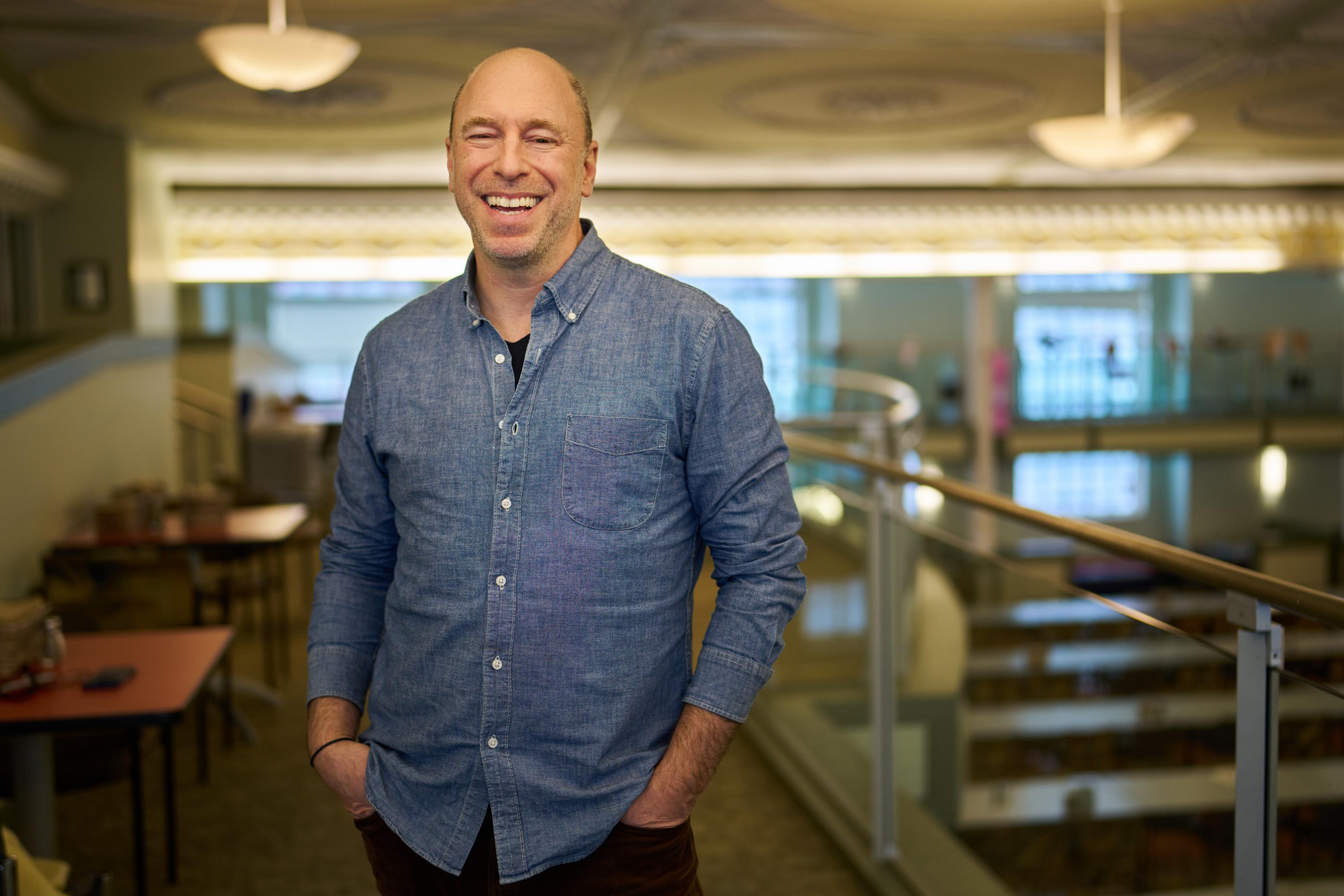 A smiling person wearing a denim shirt stands with their hands in their pants pockets in an indoor atrium with big windows.