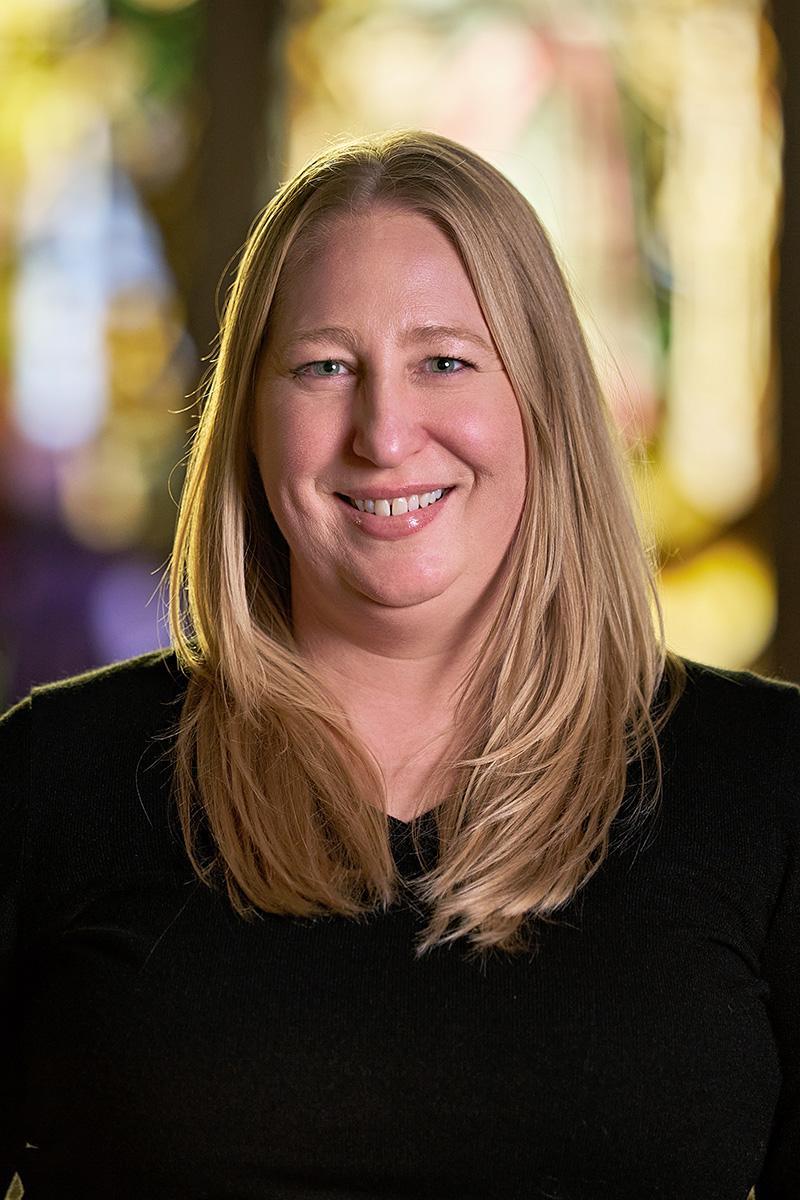 Alicia Atwood smiling with long blonde hair wearing a black top, against a softly blurred, colorful background.