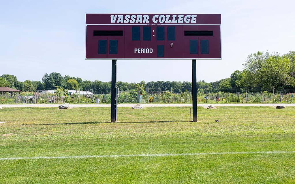 New Vassar College scoreboard on a green grass sports field, with vegetable gardens in the background.