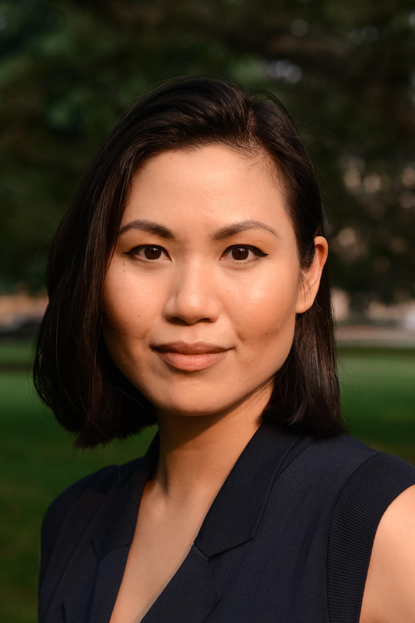  Catherine Do Tan with a short dark bob haircut wearing a sleeveless dark blazer, outdoors with greenery softly blurred in the background.