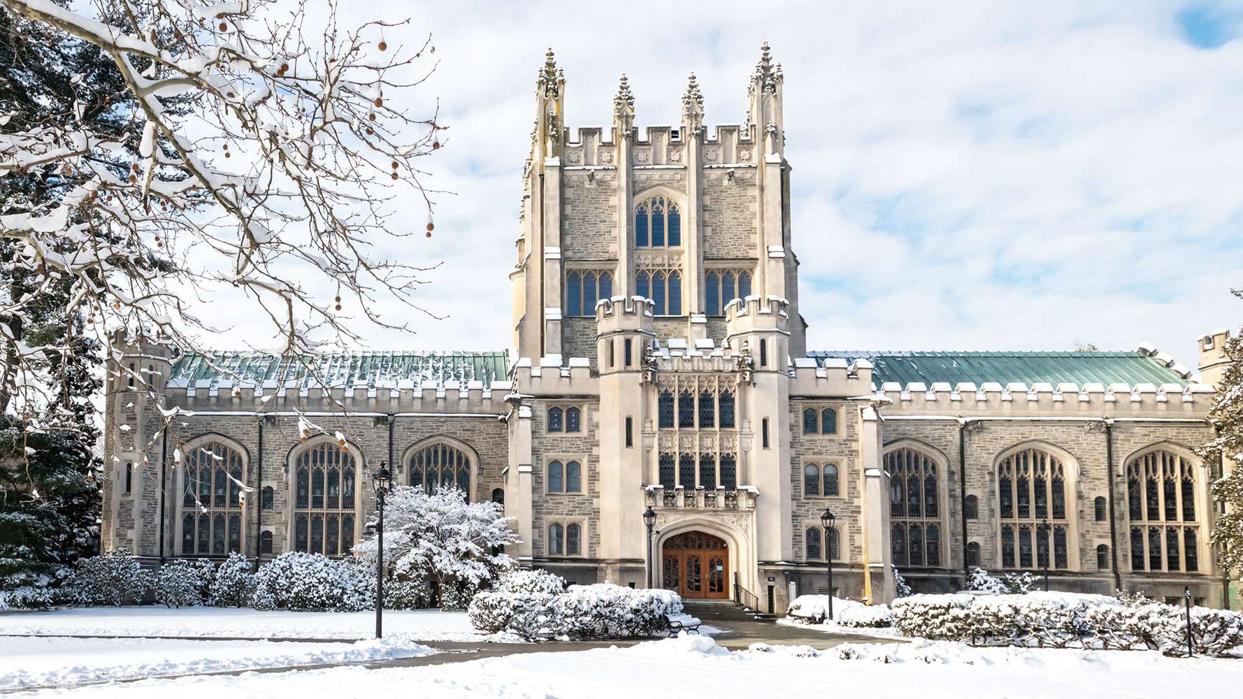 Winter view of a Gothic-style stone campus building with a tall central tower, arched windows, and a green roof, surrounded by snow-covered trees and shrubs under a partly cloudy sky.